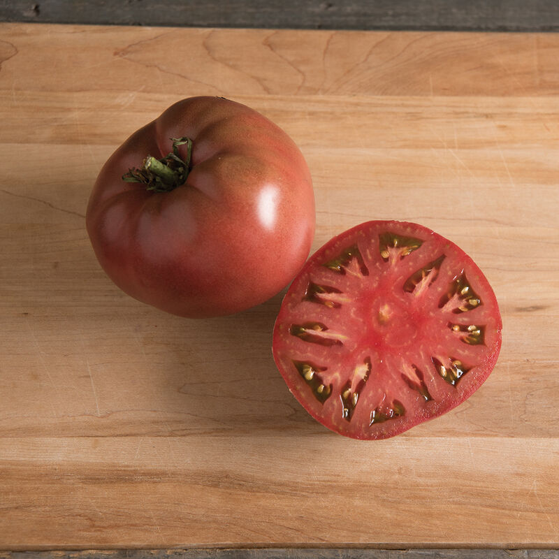 Large mature Carbon tomato fruits with dark-red to purple flesh, one whole and one sliced half on a wooden cutting board.