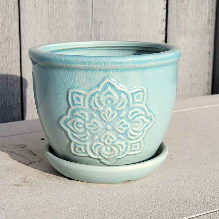 A small rounded planter with built-in saucer; the side facing the camera is decorated with mandala-style medallion element, and the planter is finished with a pale, sky blue glaze.