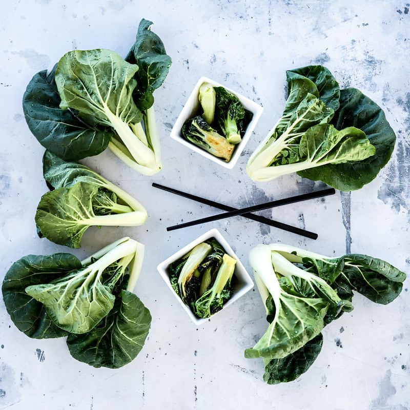 A photo of several heads of Baby Milk Bok Choy surrounding a few dishes of sauteed bok choy and a pair of chopsticks.