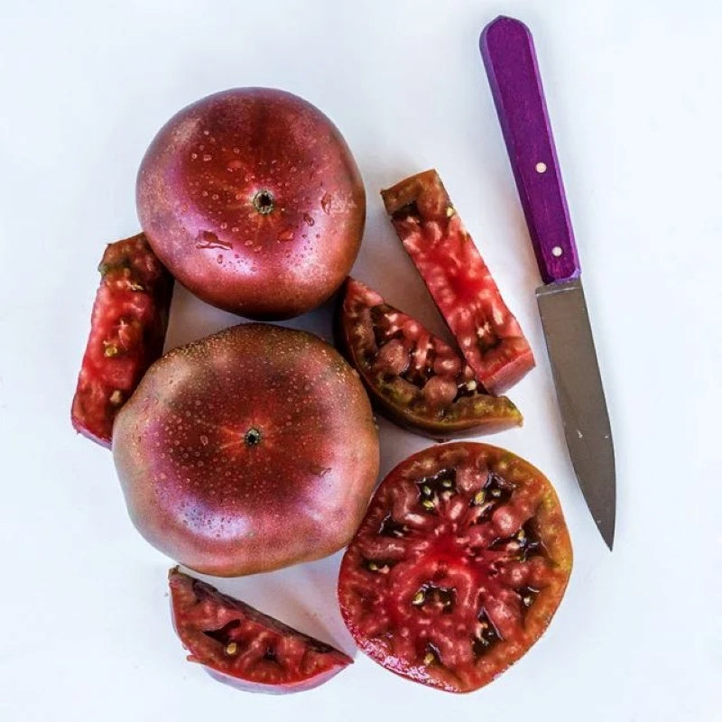 A photo of several whole and sliced Black Krim tomatoes on a white background, near a purple-handled knife.