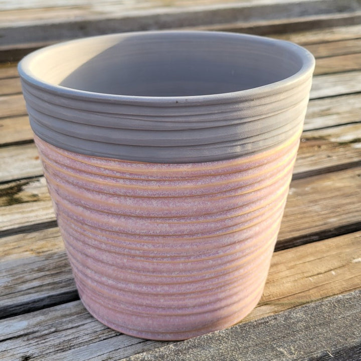 A round pot with angled walls, decorated with a carved spiral design and finished with a heathered pink and matte gray glaze combo.