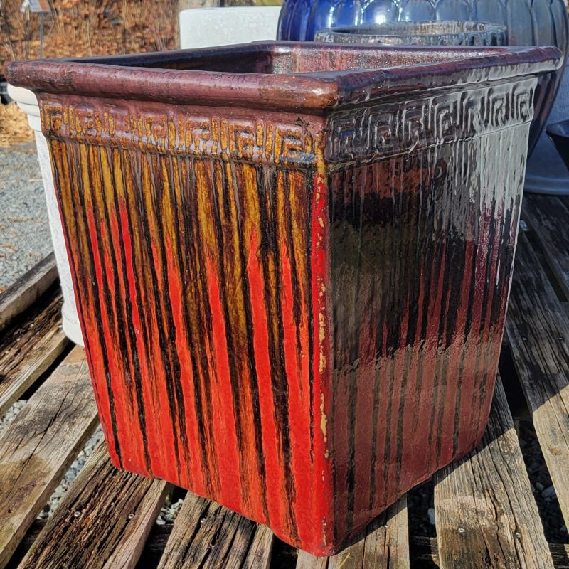 A square planter with angled walls and a pronounced lip; decorated with a faux Greco-Roman design and finished with a metallic red and dark brown drip glaze.