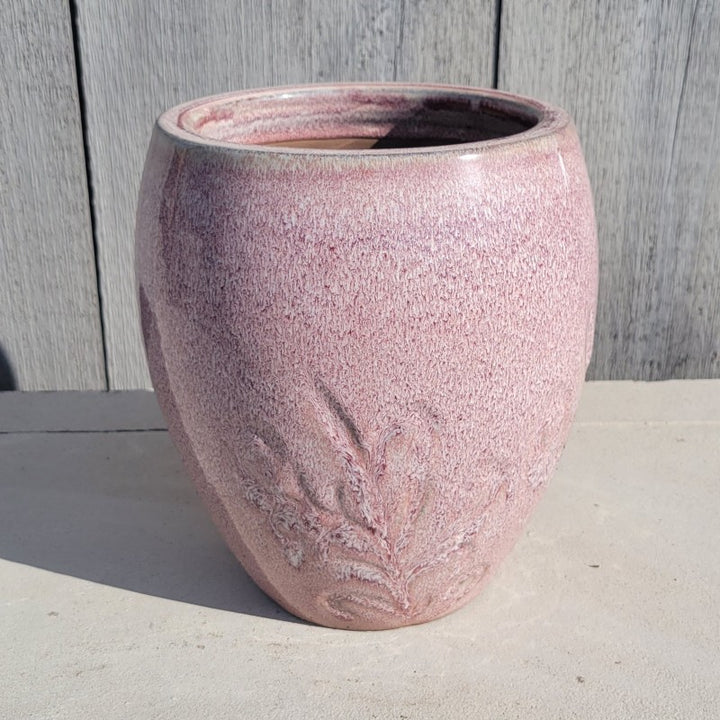 An upright, rounded, urn-style planter with a carved botanical design around the base; finished with a sunrise pink heathered glaze.