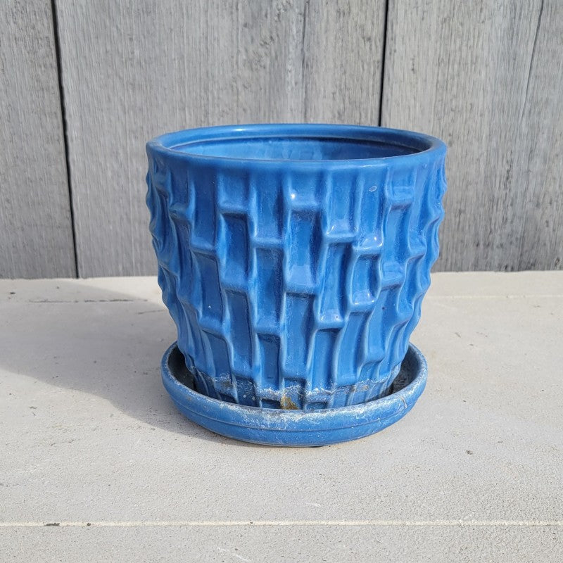 A round planter with built-in saucer and a sculpted geometric, shingle-like texture; finished in a light cobalt blue glaze.