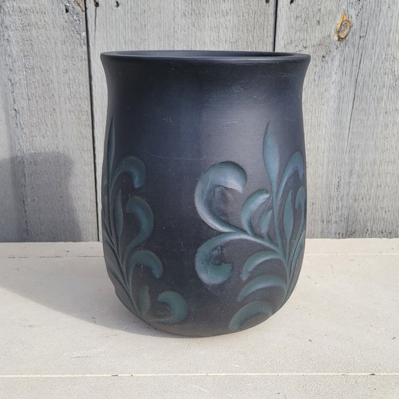 A rounded, urn-style planter with carved floral/fleur-de-lis elements; finished with matte blue-green floral elements that stand out against a field of matte black glaze.