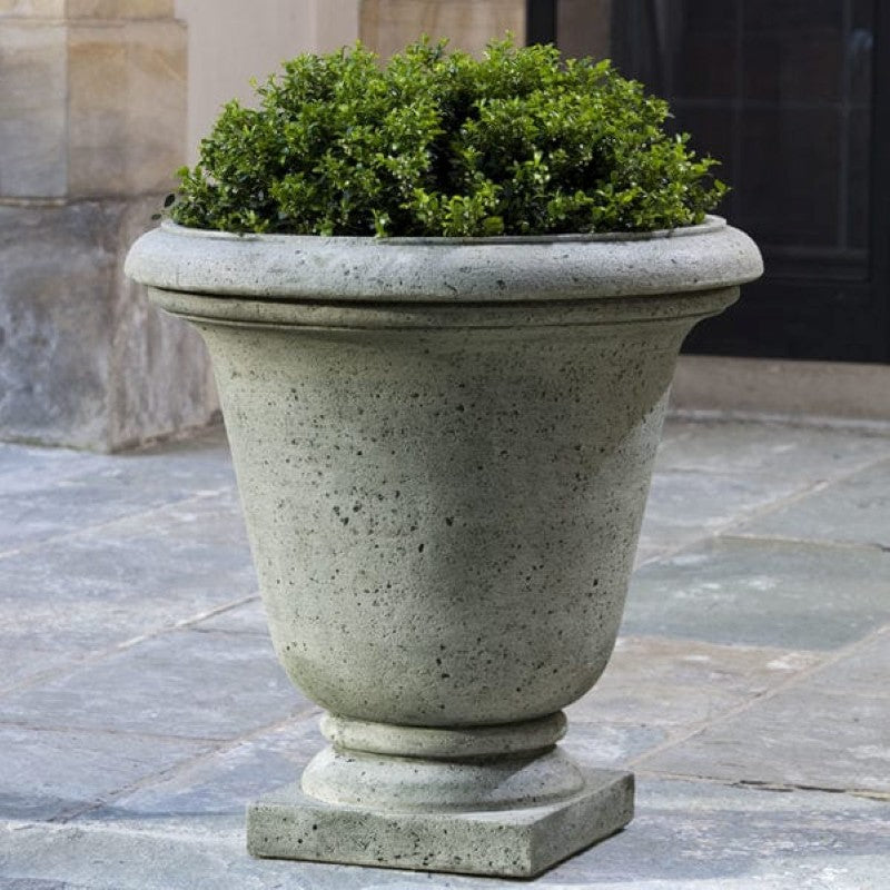 A large, cast-stone Hampton Rustic Urn with an Aged Limestone finish; placed on a paving stone patio.