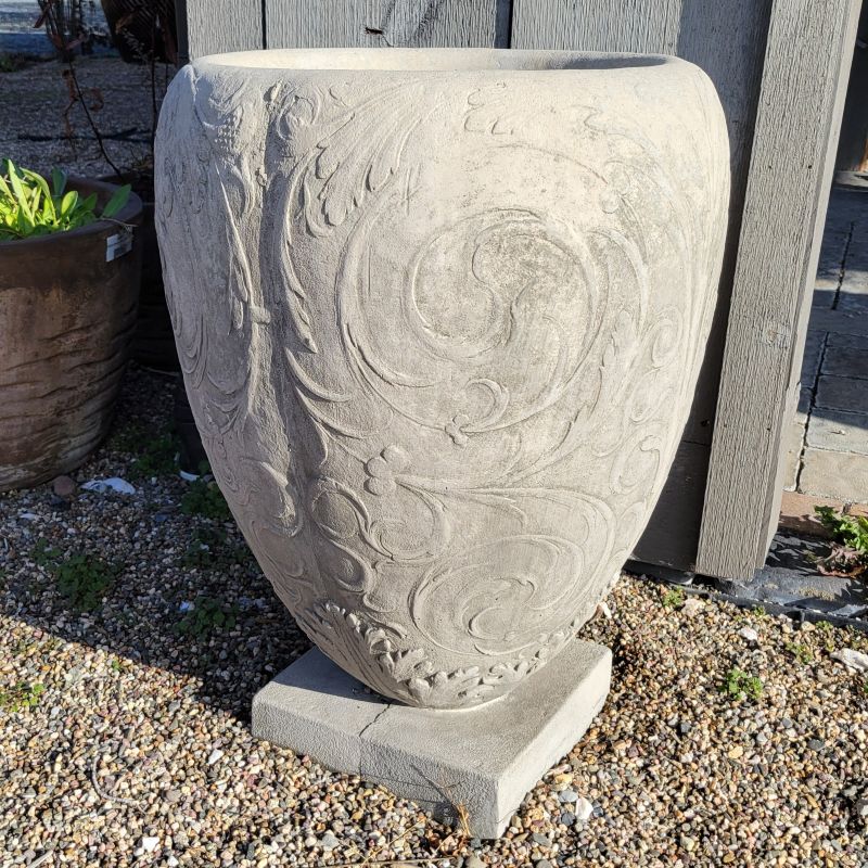 An upright, egg-shaped cast stone Longwood Arabesque Urn planter on a square base; the planter is plain, stone gray and is designed with bas-relief arabesque botanical elements.