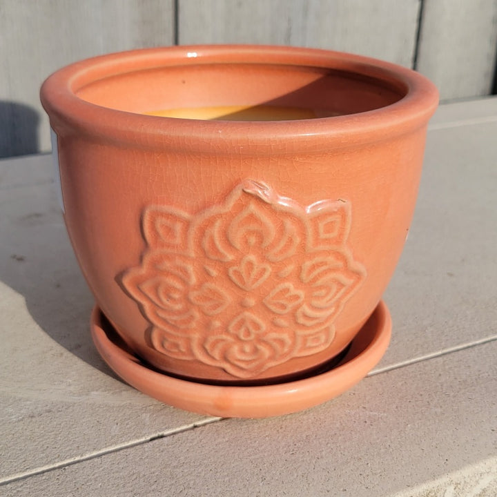 A small rounded planter with built-in saucer; the side facing the camera is decorated with mandala-style medallion element, and the planter is finished with an orange-pink glaze.